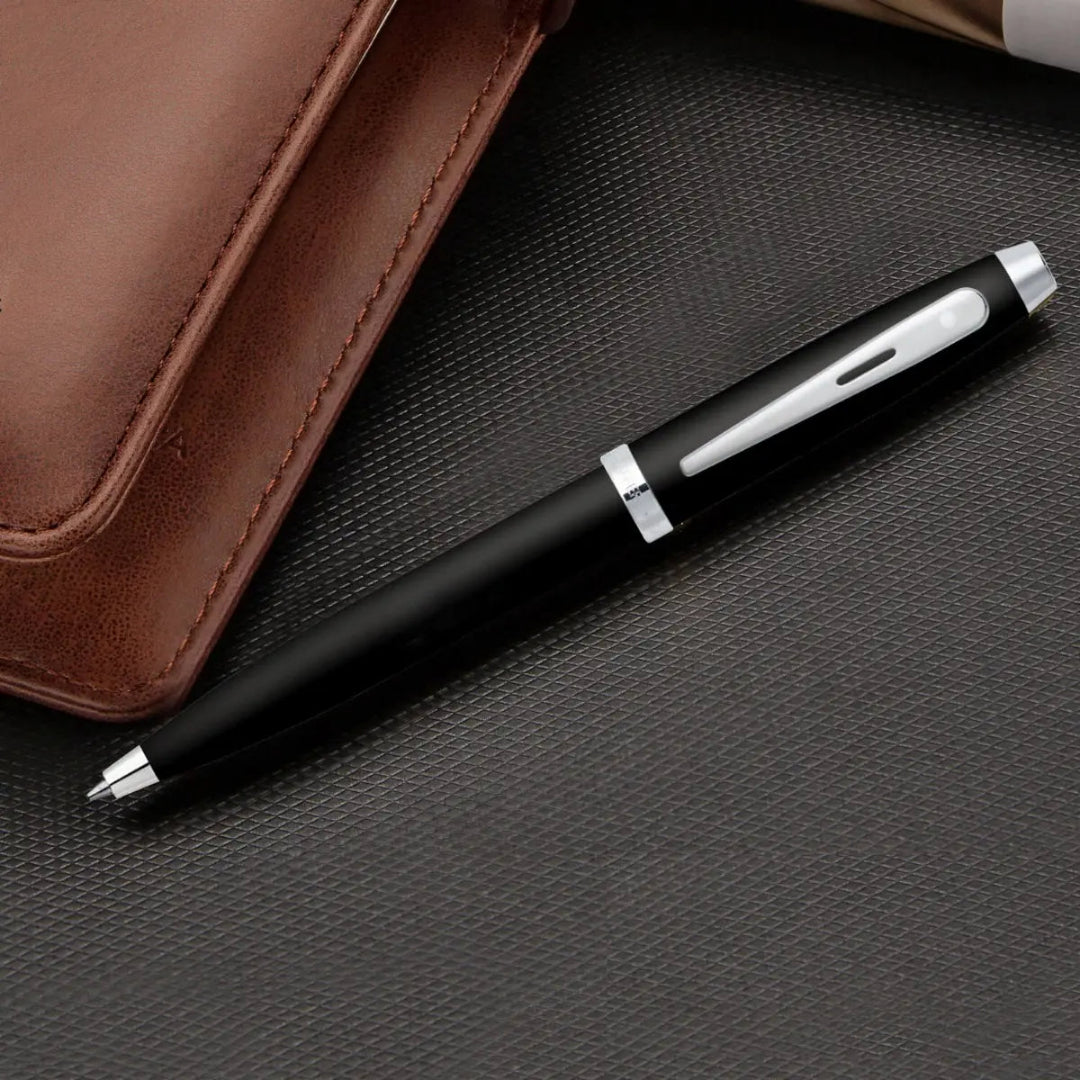 Sheaffer 100 Glossy Black Ballpoint Pen With Chrome-Plated Trims on a textured surface beside a brown leather notebook