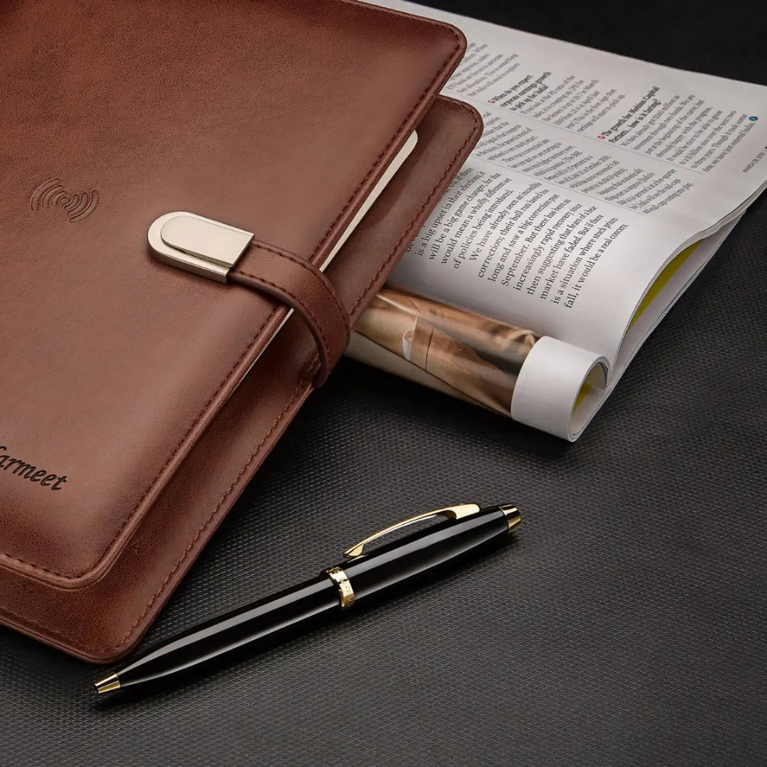 Sheaffer 100 Glossy Black Ballpoint Pen With Gold Trims beside a brown leather notebook