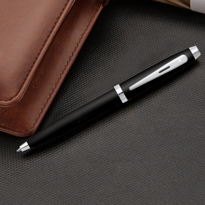 Sheaffer 100 Matte Black Ballpoint Pen With Chrome Trims on a textured surface beside a brown leather notebook